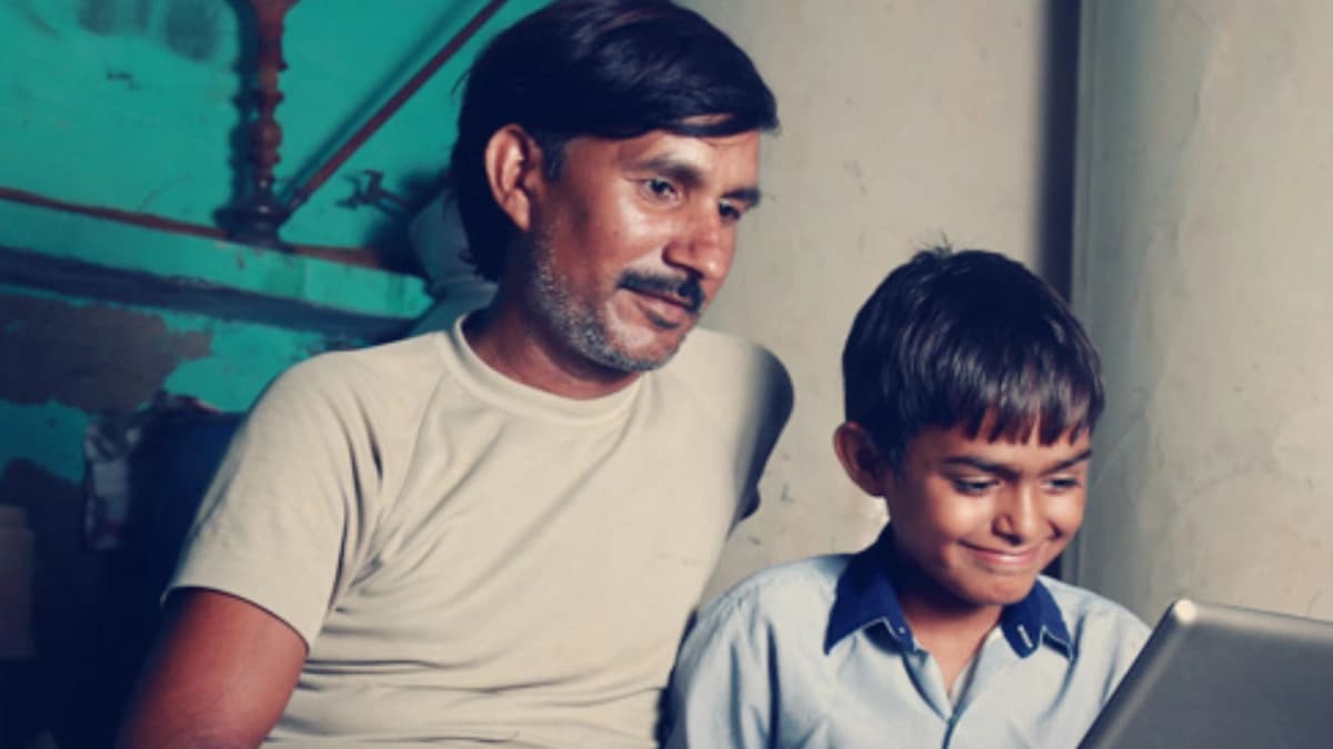 Father and son look at a laptop on the son's lap. They are in a room with tan and blue walls.