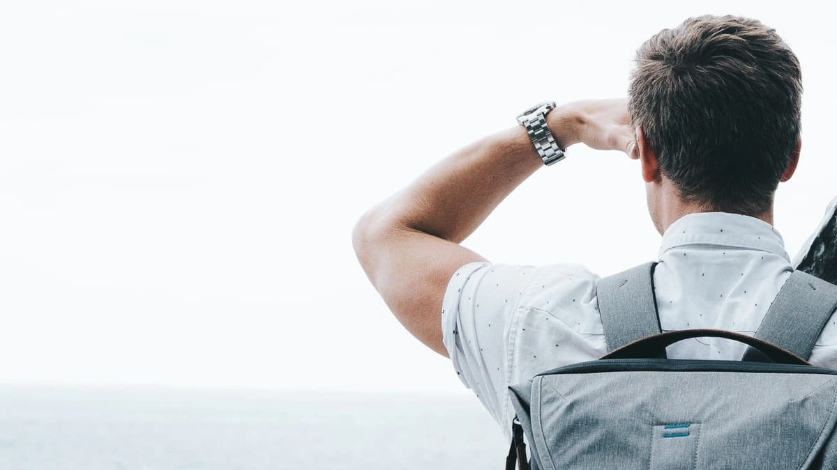 View from behind of a man with a backpack looking out into the horizon of an ocean with his arm raised and hand shading his eyes