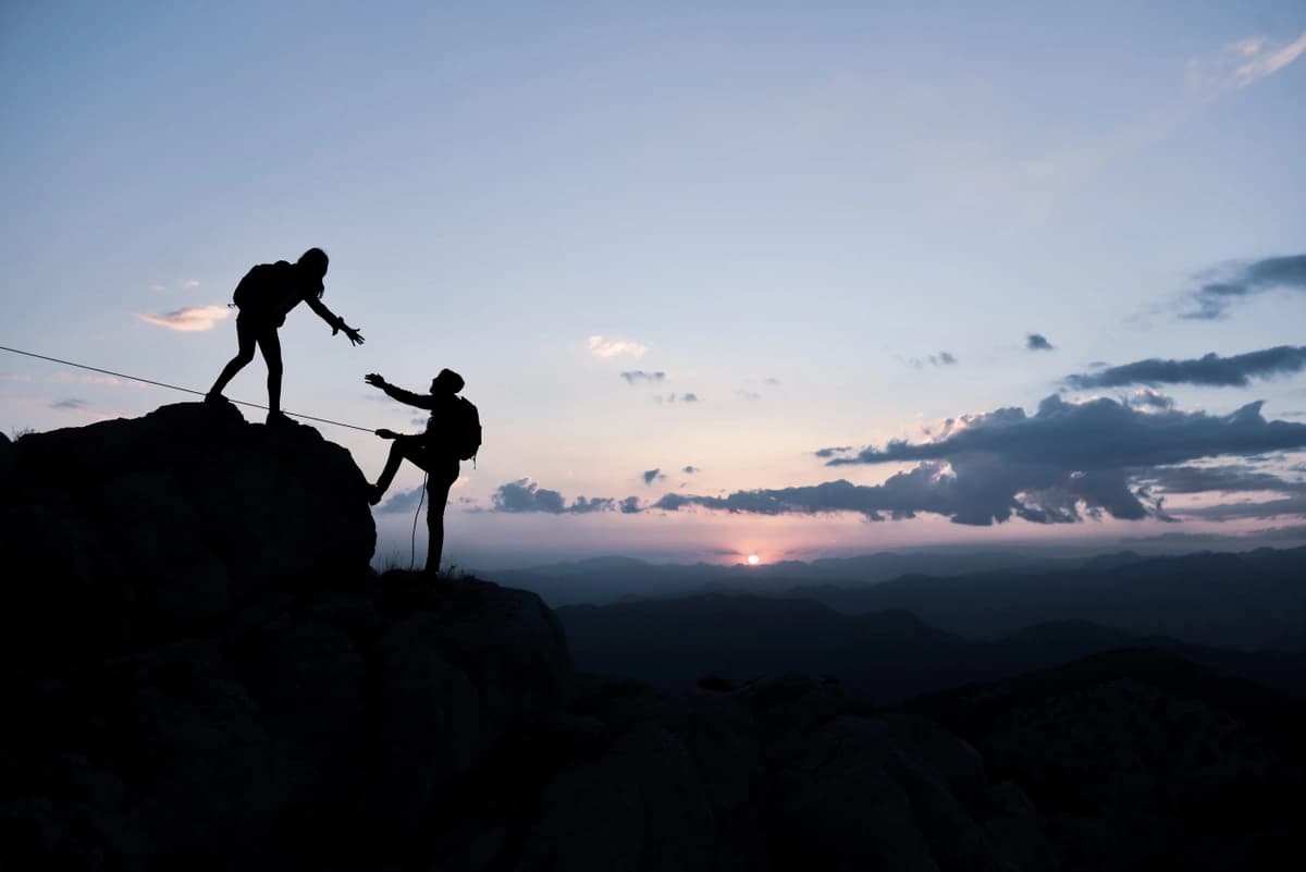 silhouette of one hiker holding hand out another climbing a rock